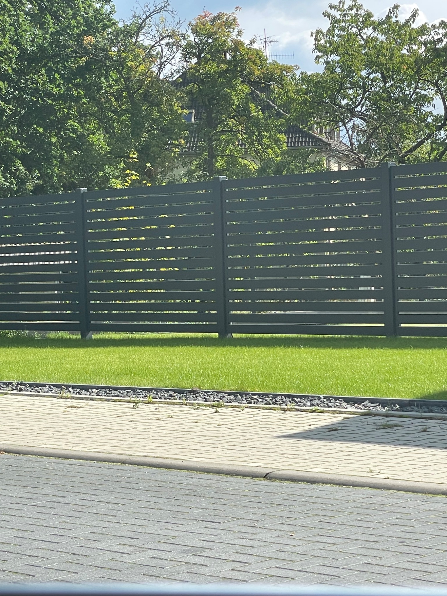 Nowoczesne, poziome, szare ogrodzenie z paneli na tle zielonego trawnika i drzew, widoczne z poziomu kostki brukowej.