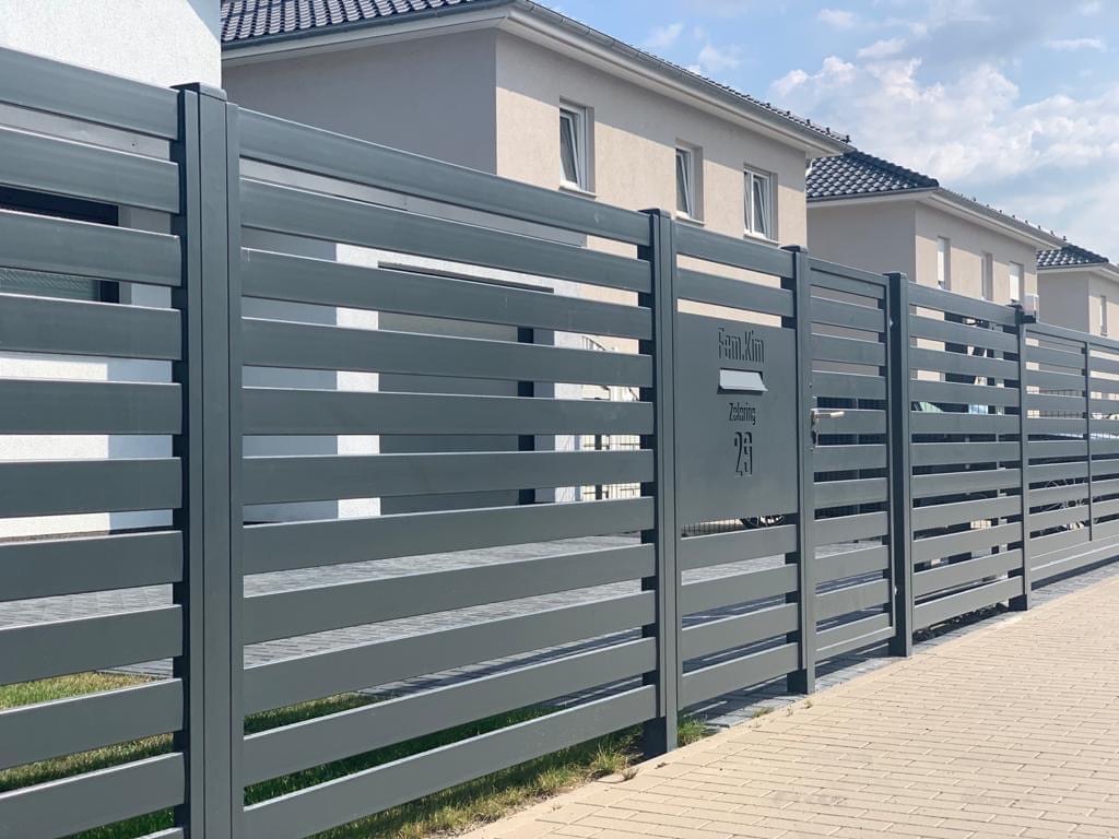 Nowoczesne, poziome ogrodzenie panelowe w kolorze grafitowym z wbudowaną skrzynką na listy i numerem domu, na tle szeregu domów jednorodzinnych.