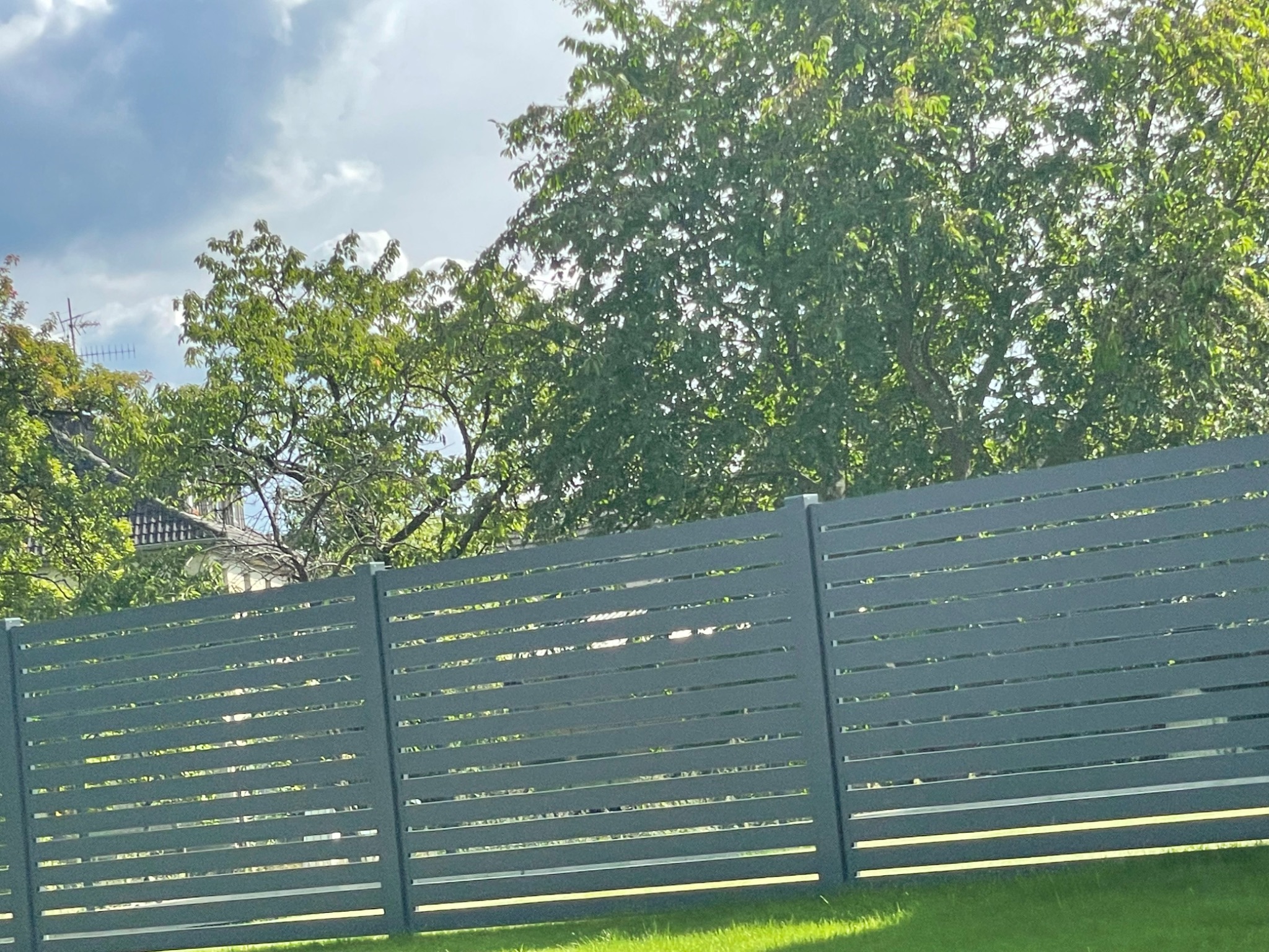 Nowoczesne, poziome ogrodzenie z szarych paneli kompozytowych na tle zielonej trawy i drzew, widoczne fragmenty domu w oddali.