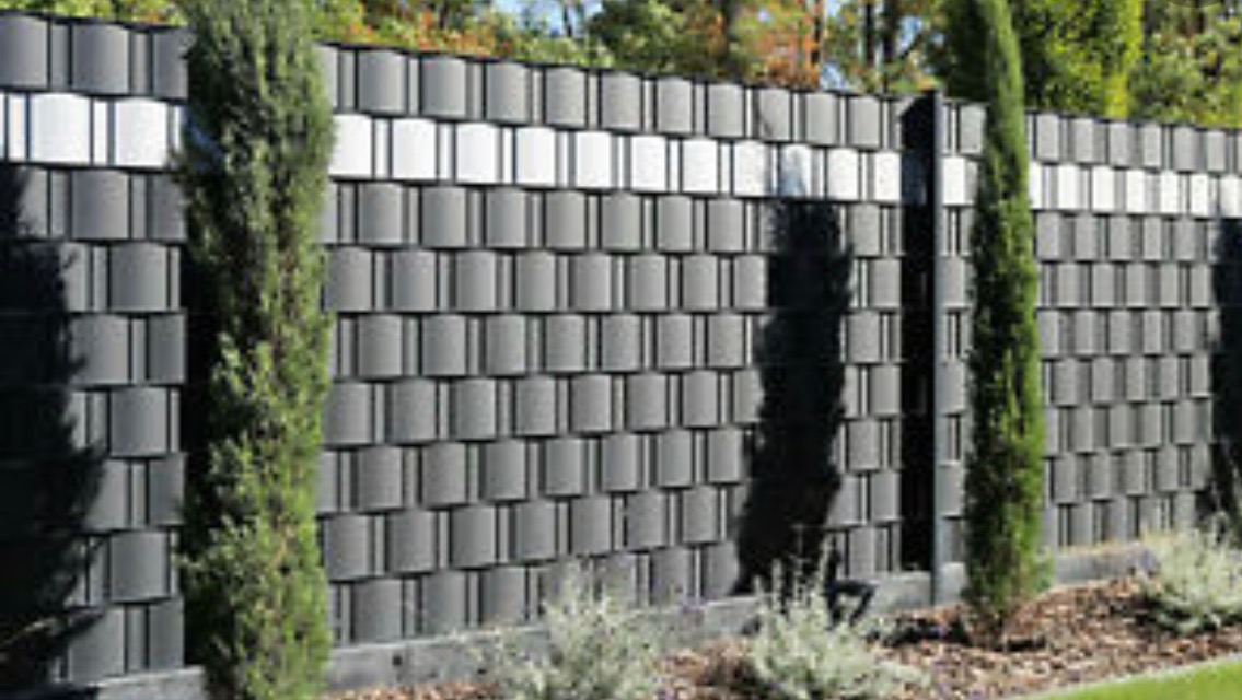 Szary płot z paneli kompozytowych z poziomymi białymi akcentami, oddzielający posesję od zieleni drzew i krzewów.