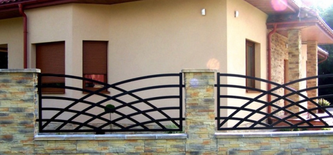 Kute ogrodzenie z czarnymi, łukowato wygiętymi przęsłami, osadzone na kamiennych słupkach i podmurówce, na tle beżowego domu z brązowymi roletami.