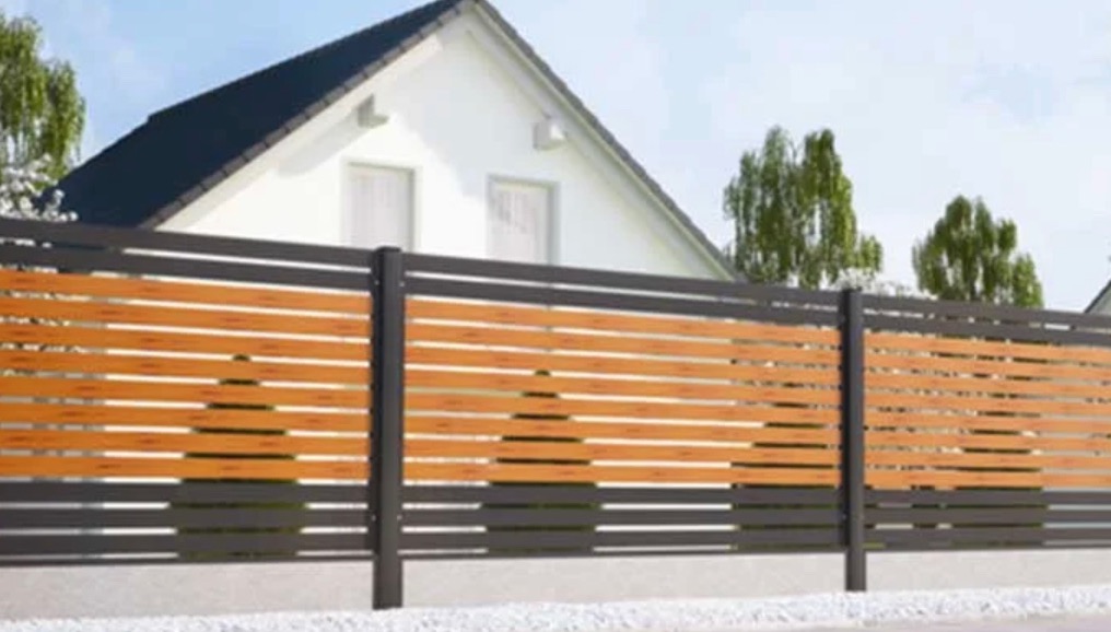 Nowoczesne ogrodzenie poziome z drewnianymi panelami w odcieniach brązu i grafitowymi słupkami na tle białego domu z ciemnym dachem.