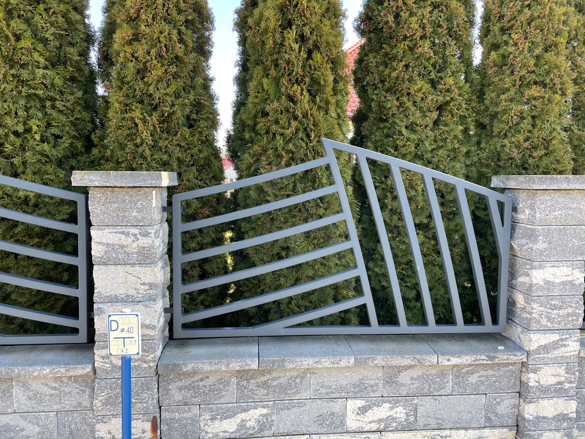 Nowoczesne ogrodzenie z szarego metalu i kamienia betonowego na tle wysokich, zielonych tuj.