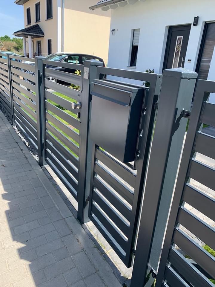 Nowoczesna, antracytowa brama wjazdowa z poziomymi panelami, zintegrowana ze skrzynką na listy, na tle jasnego domu jednorodzinnego i brukowanego chodnika.