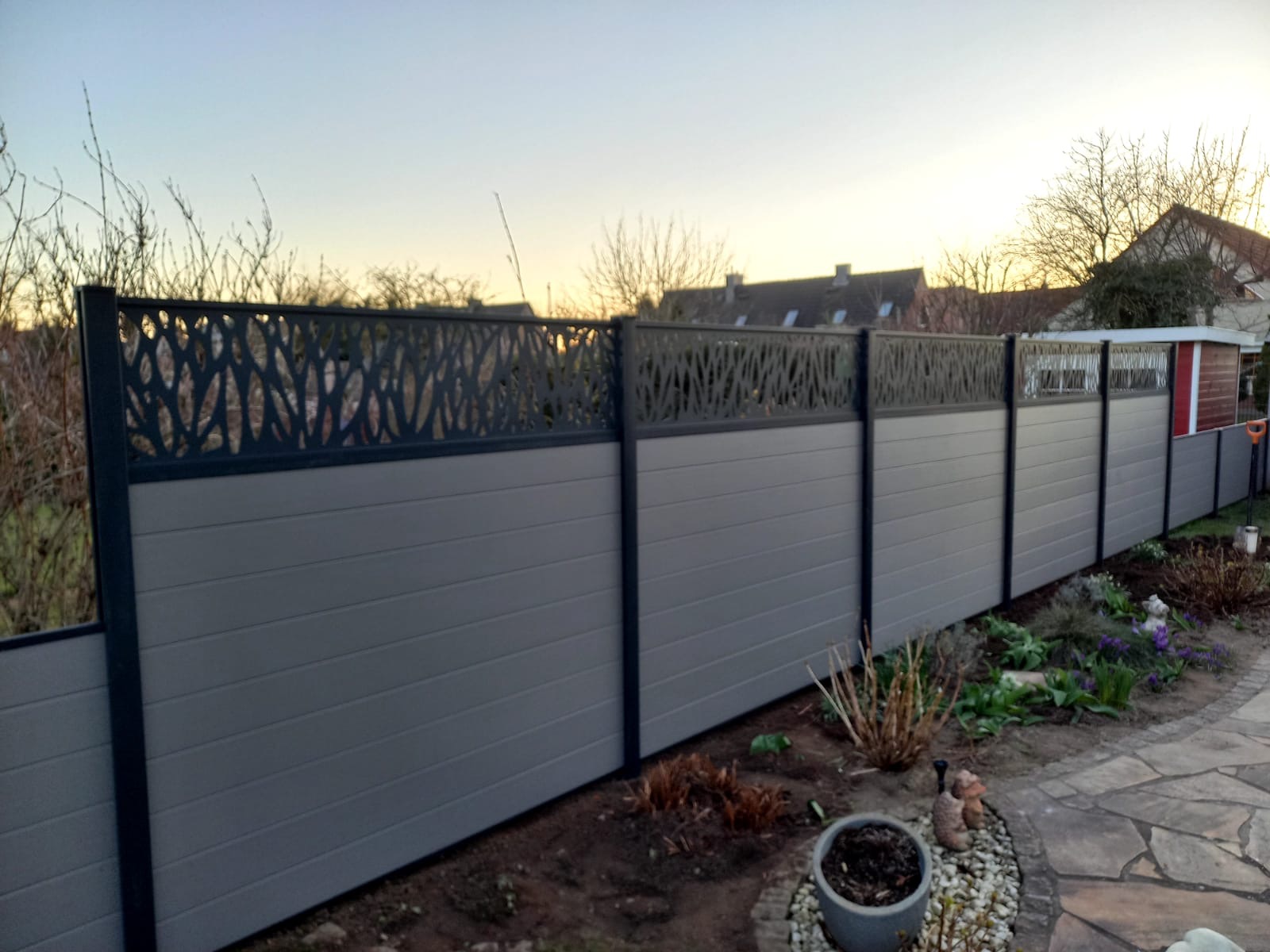 Nowoczesne ogrodzenie z szarych paneli z dekoracyjnym, ażurowym wykończeniem u góry, widoczne z perspektywy ogrodu o zmierzchu.