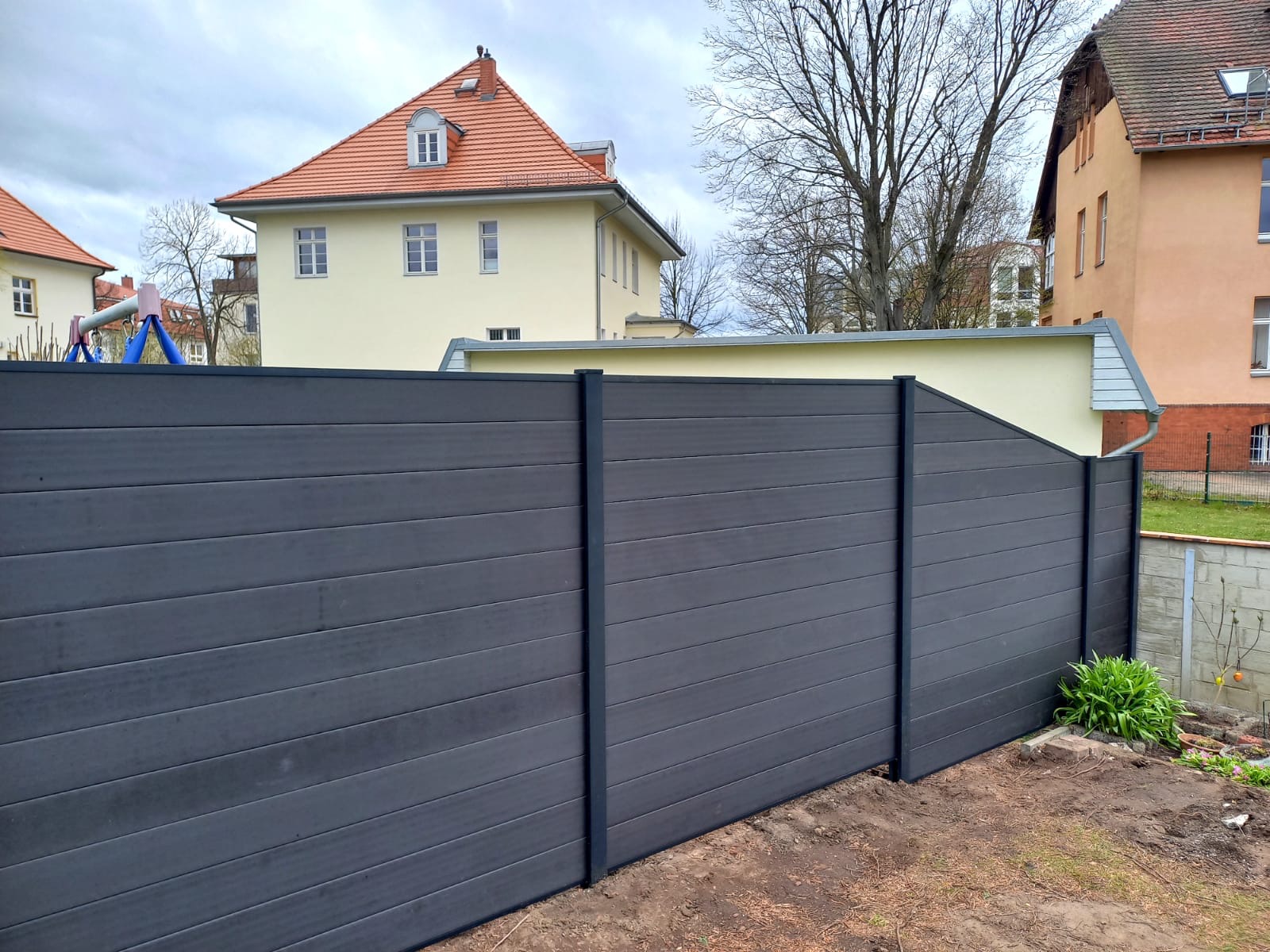 Nowoczesne ogrodzenie z kompozytu w kolorze grafitowym, z poziomymi deskami i czarnymi słupkami, na tle budynków mieszkalnych w pastelowych kolorach i pochmurnego nieba.