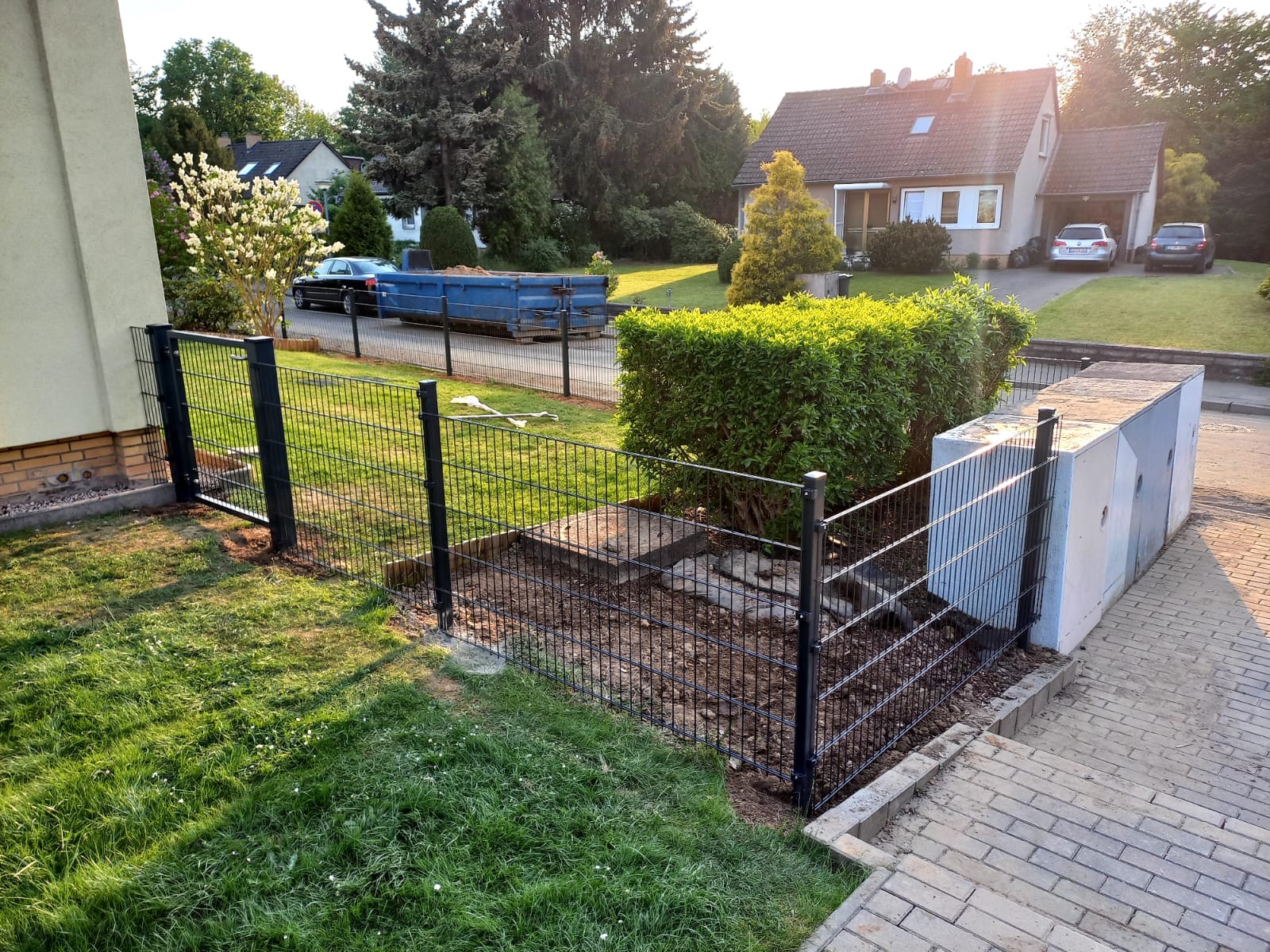 Montaż czarnego ogrodzenia panelowego na zielonej trawie, widoczny fragment posesji z domem i podjazdem, w tle kontener budowlany.