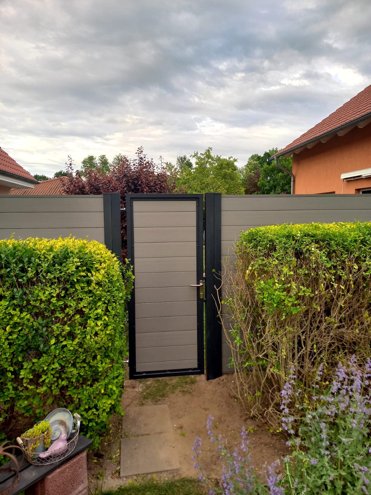 Nowoczesna furtka z paneli kompozytowych w kolorze szaro-beżowym, z czarną ramą, wkomponowana w ogrodzenie panelowe, otoczona zielenią krzewów i kwiatów.