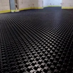 Czarna, rastrowa mata izolacyjna pod instalację ogrzewania podłogowego, rozłożona w surowym pomieszczeniu z betonowymi ścianami przygotowanymi do dalszych prac wykończeniowych.