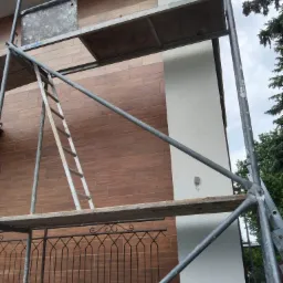 Fasada budynku z wykończeniem imitującym drewno, częściowo pokryta rusztowaniem, z widocznym fragmentem białego, otynkowanego narożnika i metalowej balustrady.