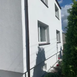 Ocieplony, pomalowany na biało narożnik domu z brązową rynną, widoczne okna z żaluzjami, fragment iglaka i czerwone róże wzdłuż fundamentu.