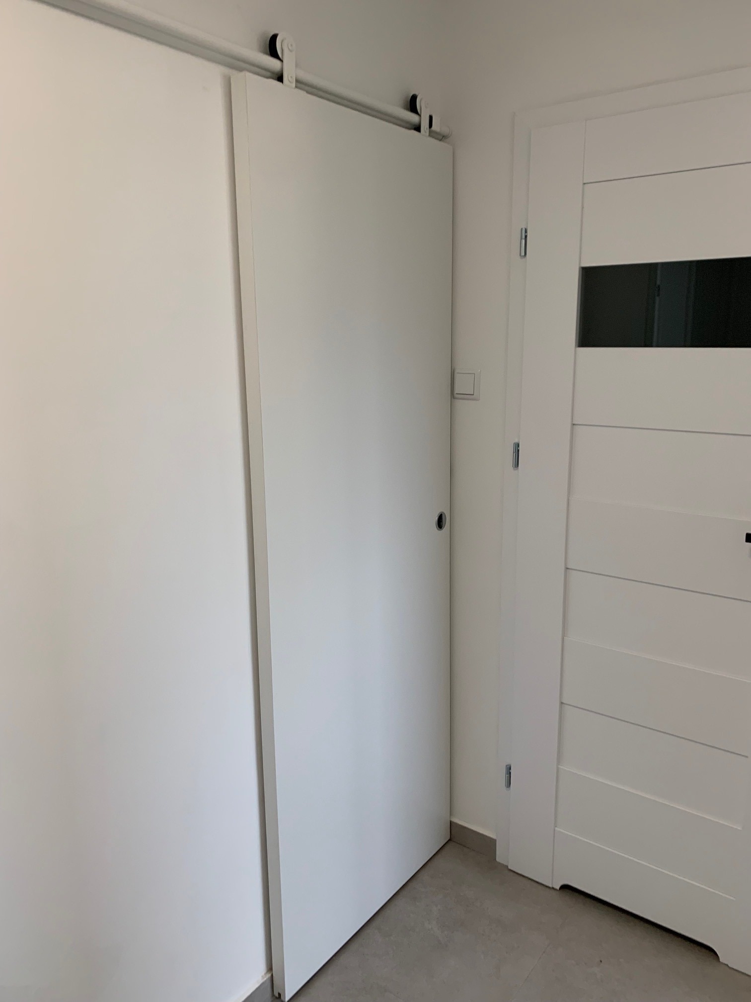Białe przesuwne drzwi zamontowane na szynie, widoczne obok białe drzwi z poziomymi panelami i czarnym okienkiem, szary podłoga.