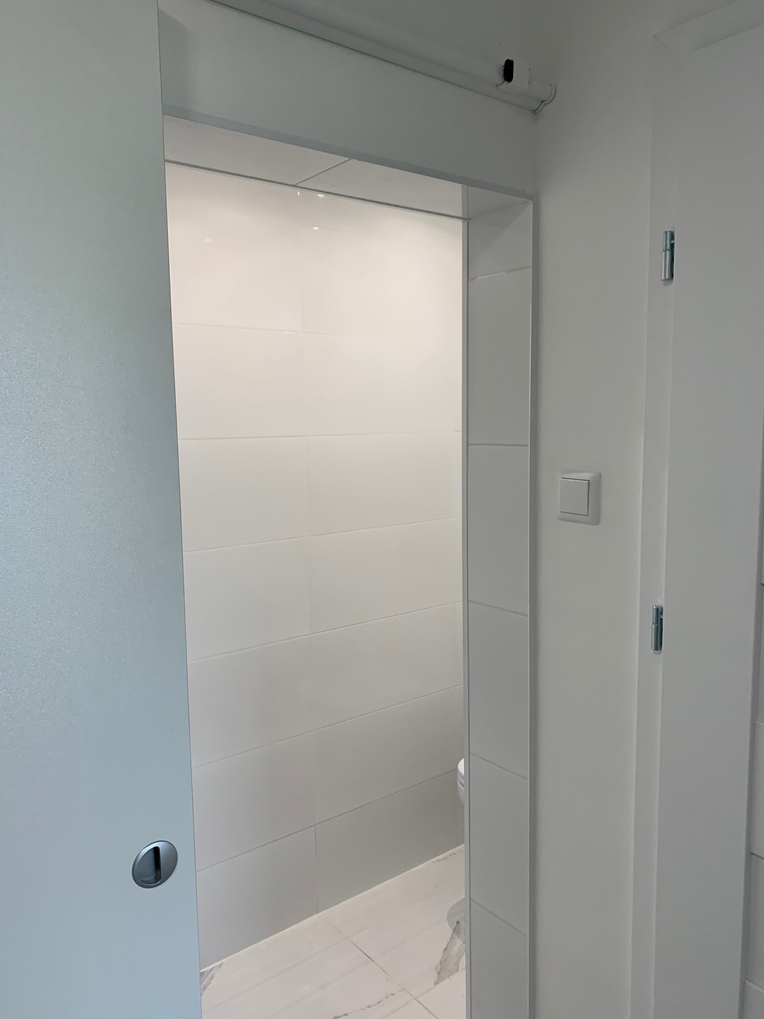 Wnętrze łazienki w minimalistycznym stylu z przesuwnymi drzwiami, białe płytki na ścianach i podłodze imitują marmur, widoczny fragment toalety.