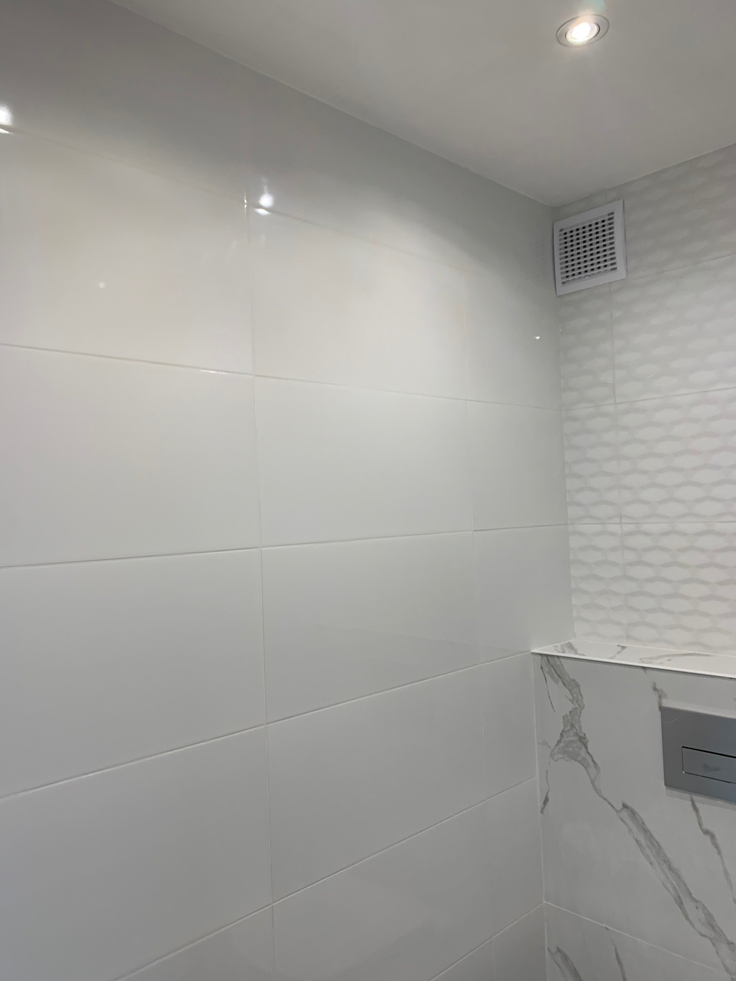 Biała, błyszcząca glazura ścienna w łazience, z dekoracyjną mozaiką i wentylatorem w rogu, oświetlona punktowym światłem.