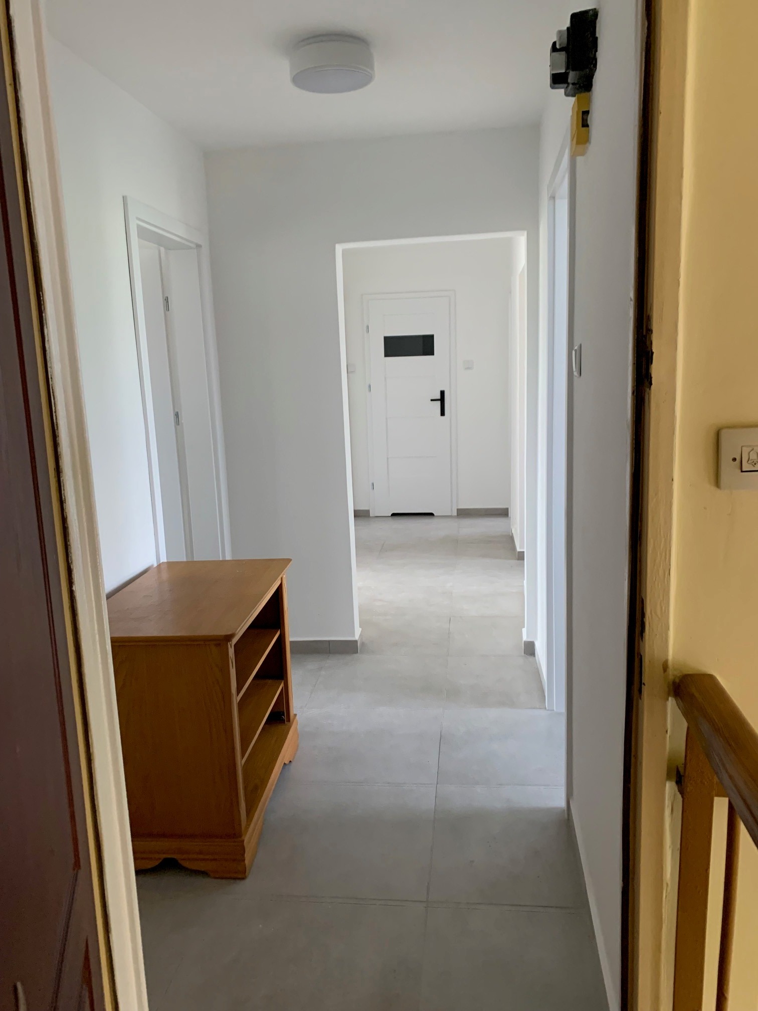 Wnętrze korytarza po remoncie z szarą podłogą, białymi ścianami i drewnianą komodą przy ścianie, widok na białe drzwi z czarną klamką na końcu korytarza.