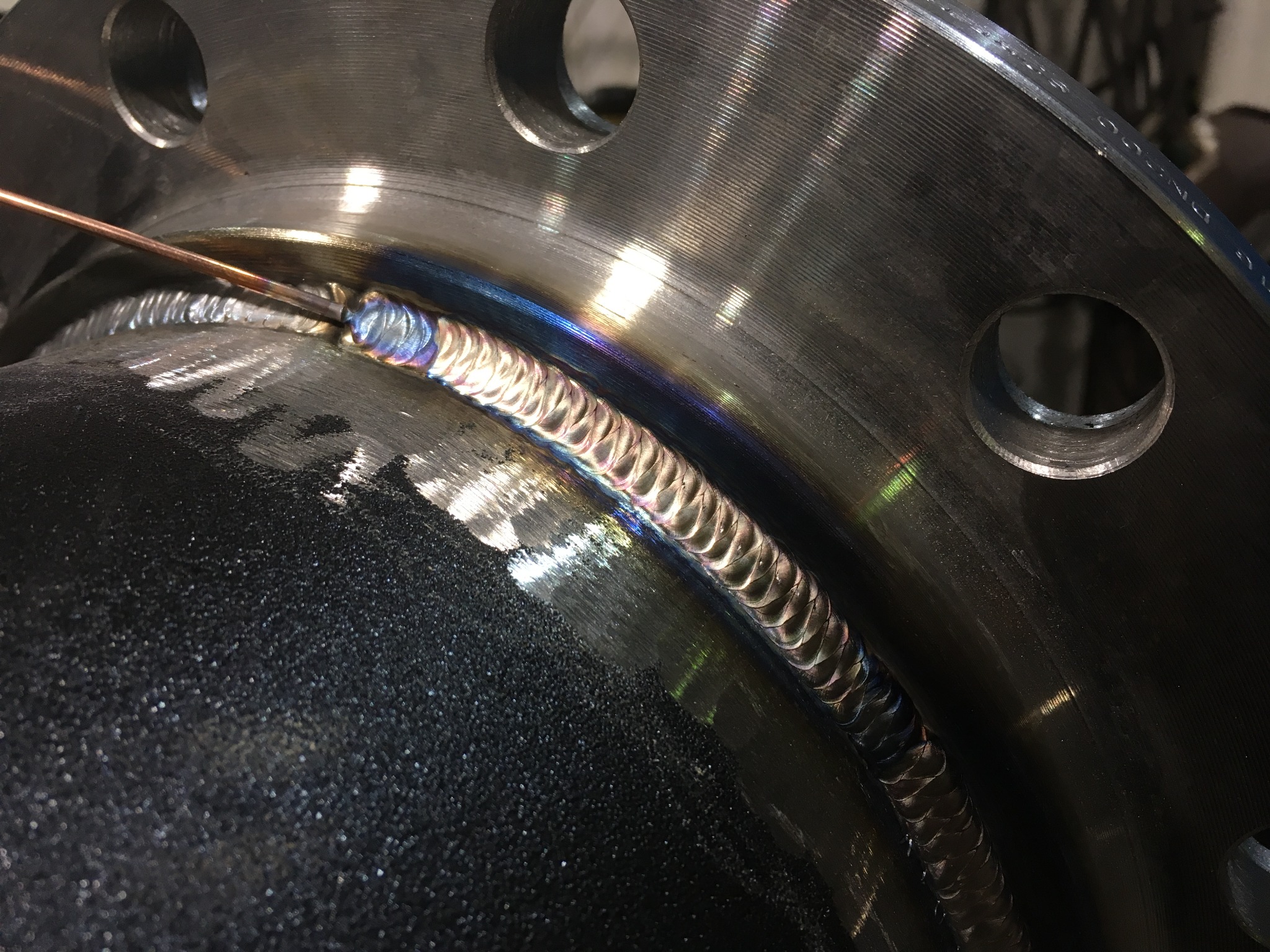 Spoina na metalowej rurze z kołnierzem, widoczne tęczowe zabarwienie metalu po spawaniu.