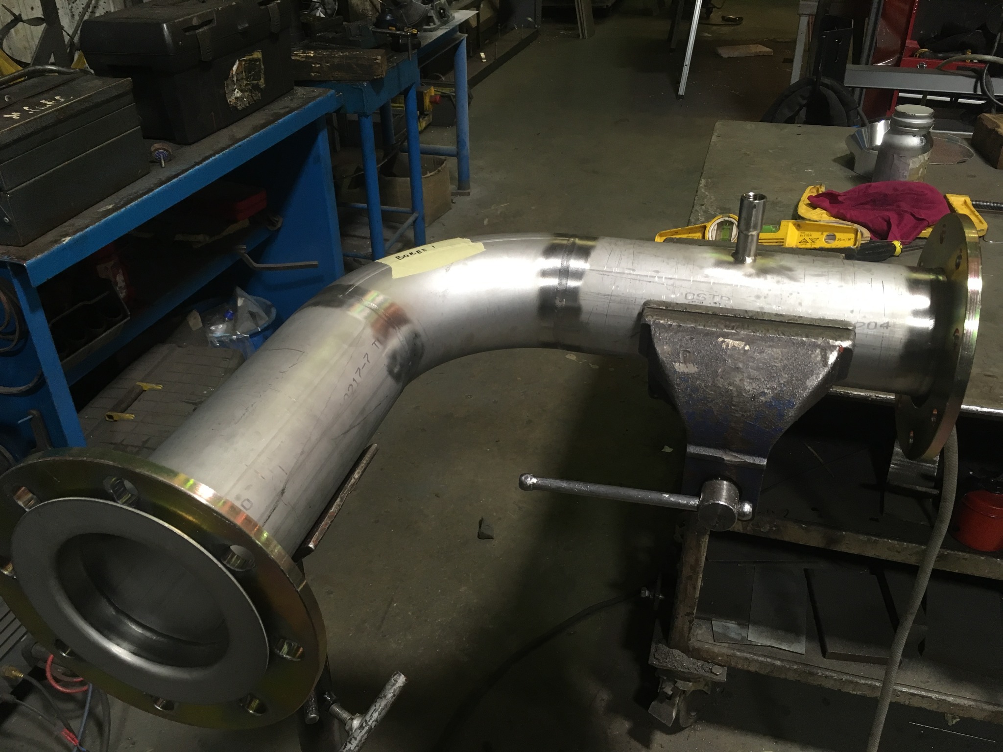 Gięty element rurociągu ze stali nierdzewnej z przyspawanym kołnierzem, zamocowany w imadle na warsztatowym stole, z widocznymi śladami po obróbce.