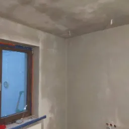 Wnętrze pokoju po nałożeniu gładzi, widoczne ślady po szpachlowaniu na ścianach i suficie, okno z brązową ramą i grzejnik pod nim, wystające kable ze ściany.