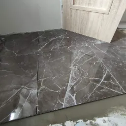 Układanie ciemnych, marmurkowych płytek podłogowych z białymi żyłkami, z użyciem krzyżyków dystansowych, widoczna surowa wylewka i fragment drzwi.