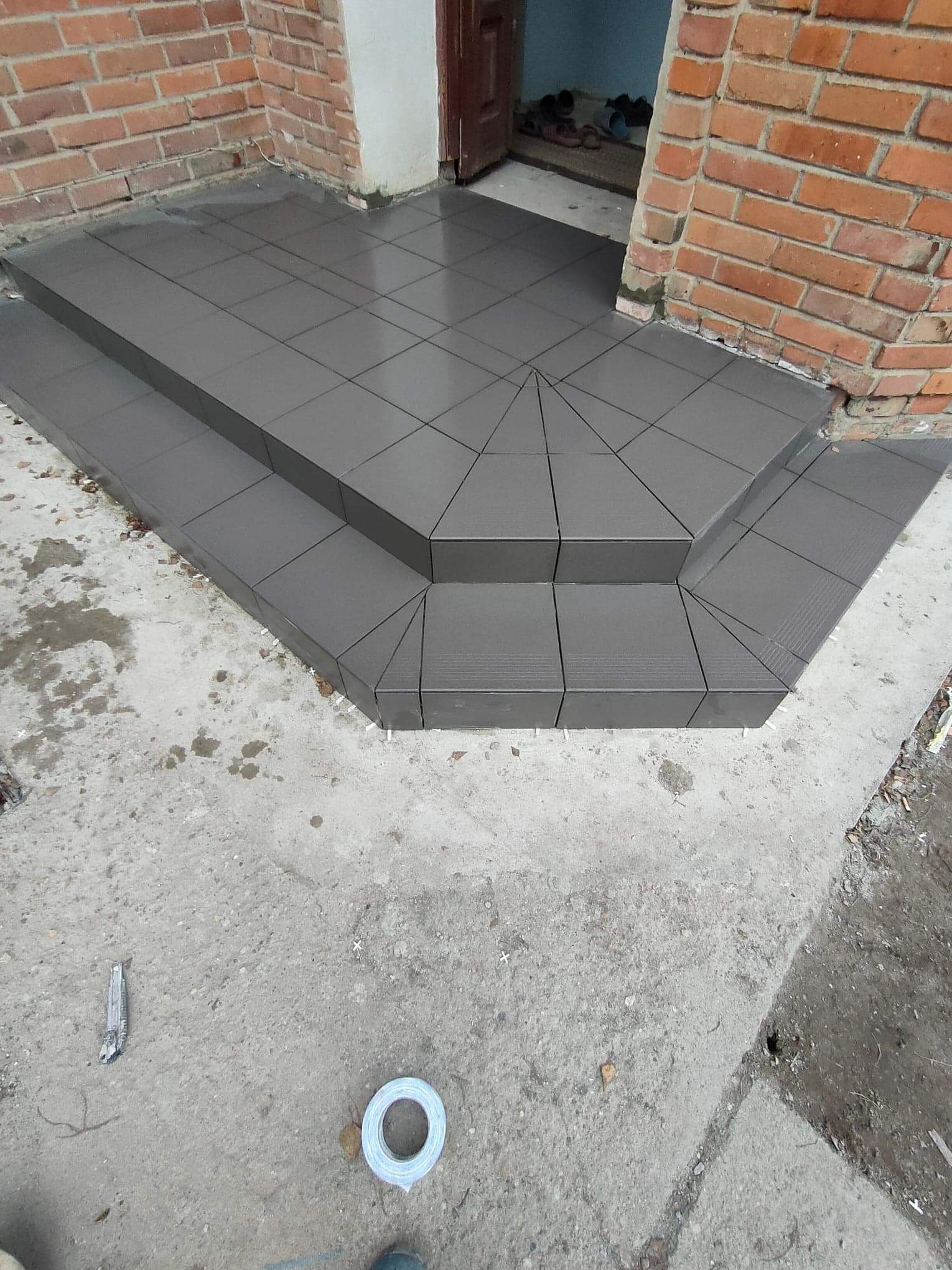 Wykończone płytkami ceramicznymi schody wejściowe do domu z ceglaną elewacją, widoczne narzędzia glazurnicze na betonowym podłożu.