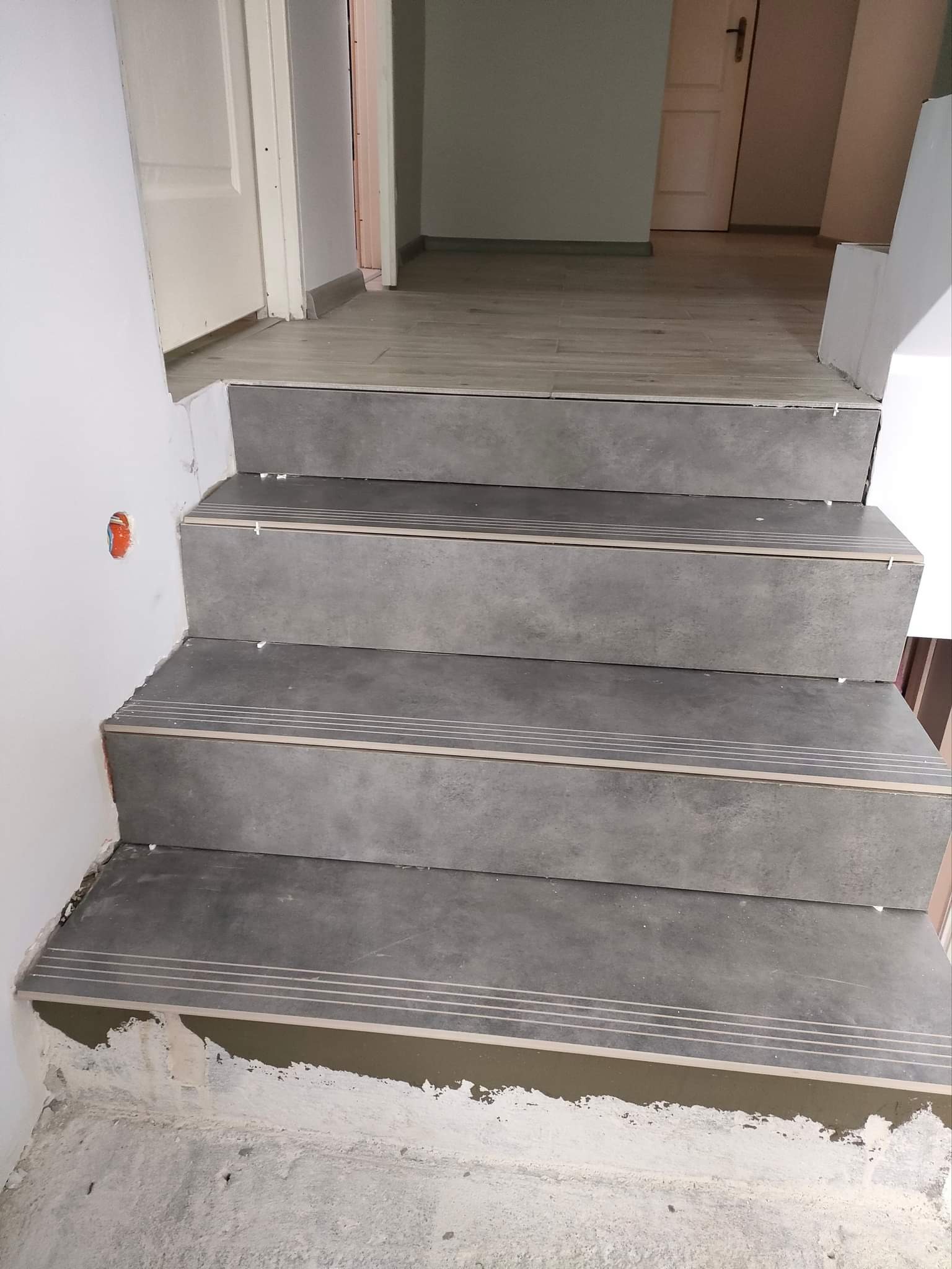 Wykończone schody z szarych płytek z metalowymi listwami antypoślizgowymi, widoczne niedociągnięcia w postaci śladów zaprawy i odkrytej instalacji elektrycznej, w tle drewnopodobna podłoga i białe...