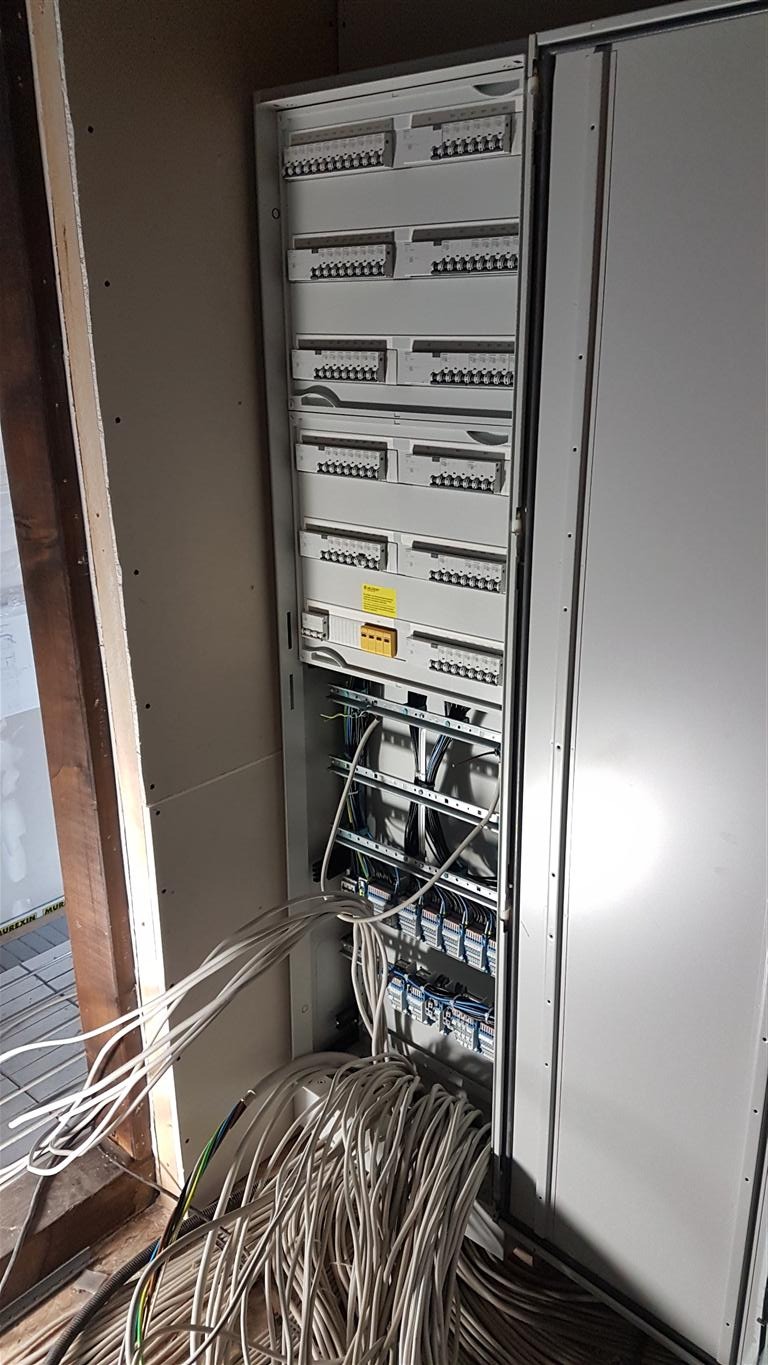 Rozdzielnica elektryczna z podłączonymi przewodami w surowym stanie, widoczne zabezpieczenia i moduły, ściany w trakcie wykończenia.