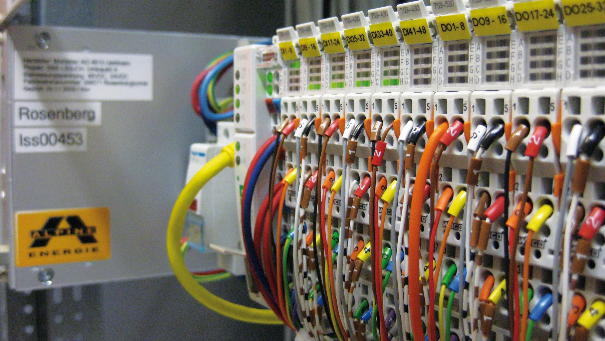 Szczegółowe zbliżenie na panel sterowniczy z kolorowymi przewodami i oznaczeniami, widoczna tabliczka znamionowa Rosenberg i logo Alpins Energie.