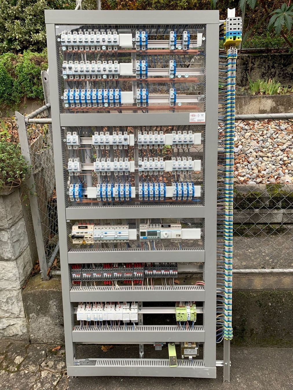 Szara szafa sterownicza z licznymi zabezpieczeniami, przekaźnikami i okablowaniem, widoczna na zewnątrz budynku przy siatce ogrodzeniowej.
