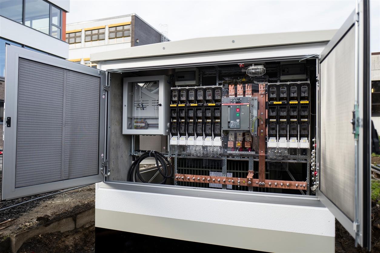 Otwarta szafa elektryczna na zewnątrz budynku, widoczne zabezpieczenia, kable i szyny miedziane, z elementami instalacji wewnątrz.