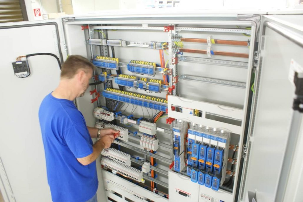 Elektryk w niebieskiej koszulce, z miernikiem w dłoni, pracuje nad rozbudowaną szafą sterowniczą z licznymi zabezpieczeniami, przekaźnikami i okablowaniem.