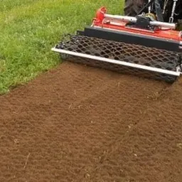 Czerwony traktor z czarnym wałem siatkowym wyrównuje świeżo przekopaną ziemię, pozostawiając równą, brązową powierzchnię obok zielonego trawnika.