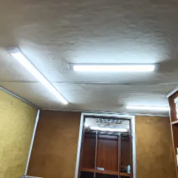 Wnętrze kampera z tapicerowanymi ścianami w odcieniach brązu i beżu, oświetlone ledowymi lampami sufitowymi, z widoczną szafą wnękową i skrzynką elektryczną.