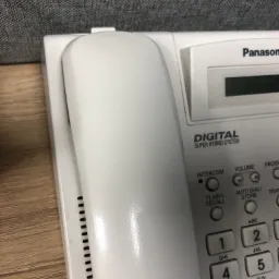 Biały telefon stacjonarny Panasonic Digital Super Hybrid System z widoczną klawiaturą numeryczną, przyciskami funkcyjnymi i wyświetlaczem, leżący na drewnianej powierzchni, z szarym tłem.