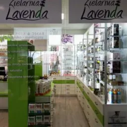 Wejście do zielarni 'Lavenda' z widocznymi półkami pełnymi produktów ziołowych i kosmetyków naturalnych, zielone akcenty kolorystyczne, widok z zewnątrz przez szklane drzwi.