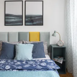 Jasna sypialnia z łóżkiem z panelowym zagłówkiem w odcieniach szarości i żółci, niebieską pościelą w białe kwiaty, turkusową narzutą, szafką nocną z lampką i zegarem oraz dwoma obrazami w czarnych...