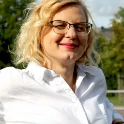 Portret uśmiechniętej kobiety w okularach i białej koszuli na tle zieleni miejskiej.