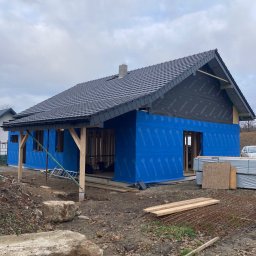 ONY DOMY SZKIELETOWE ADAM SROKA - Budowa domu szkieletowego w trakcie realizacji: widoczne drewniane elementy konstrukcyjne, niebieska membrana i czarna dachówka. Częściowo wykończony dach i ściany.