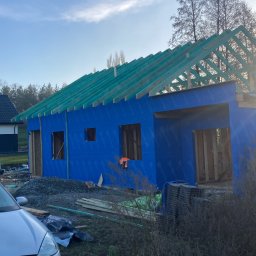 ONY DOMY SZKIELETOWE ADAM SROKA - Budowa domu szkieletowego: niebieska elewacja, drewniana konstrukcja dachu z zieloną membraną, widoczne okna i drzwi. Częściowo widoczny samochód osobowy.