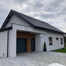 ONY DOMY SZKIELETOWE ADAM SROKA - Nowoczesny dom z garażem i drewnianymi akcentami, wykończony jasną elewacją i ciemną dachówką. Podjazd wyłożony kostką brukową, małe drzewko w donicy przy wejściu.
