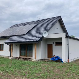 ONY DOMY SZKIELETOWE ADAM SROKA - Gotowy dom szkieletowy z panelami słonecznymi na dachu, antena satelitarna, drewniane elementy wykończeniowe, trawa i ziemia wokół budynku.