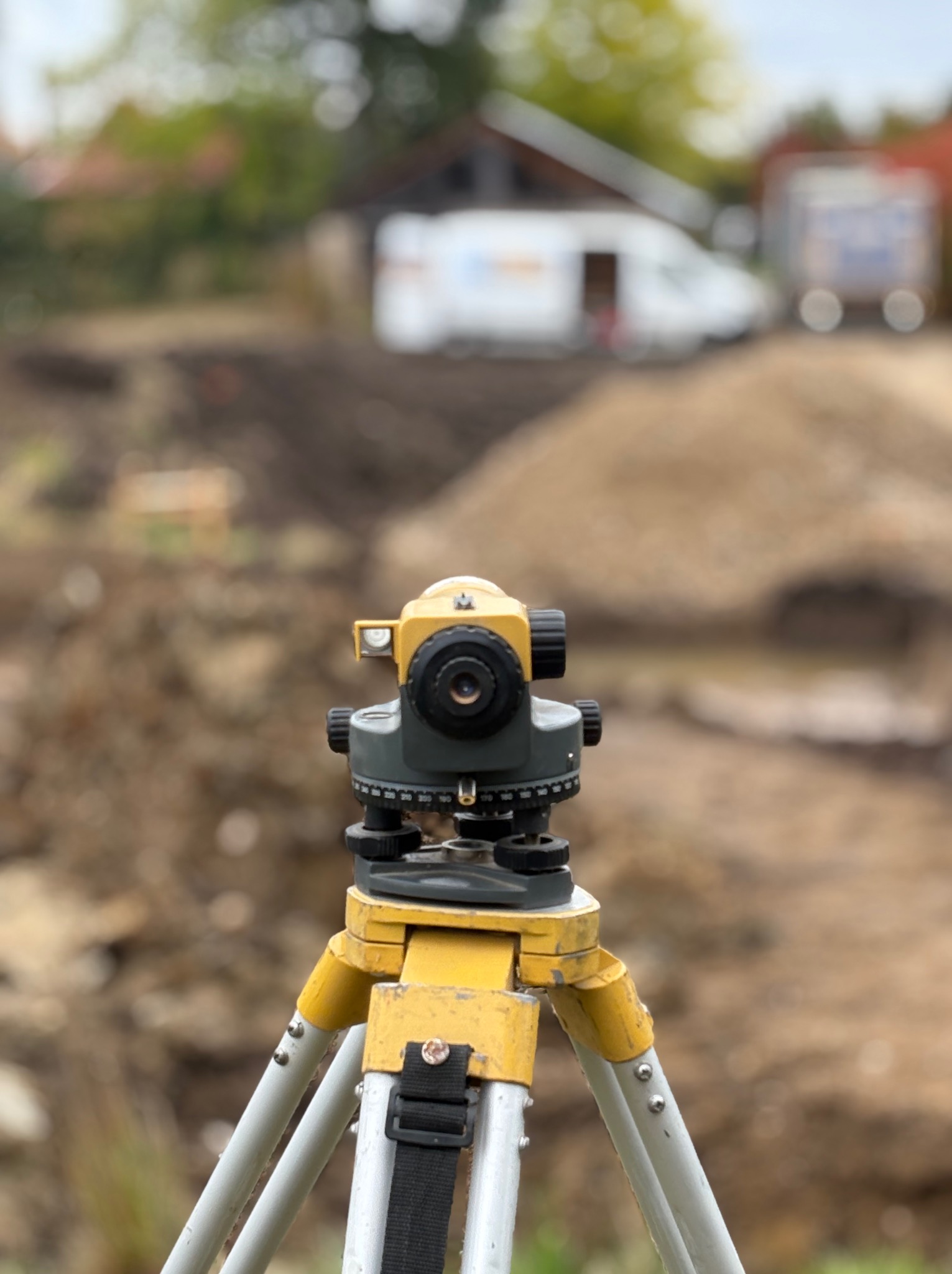 Żółty niwelator optyczny na trójnogu podczas prac ziemnych pod budowę domu szkieletowego, w tle wykop i budynek gospodarczy.