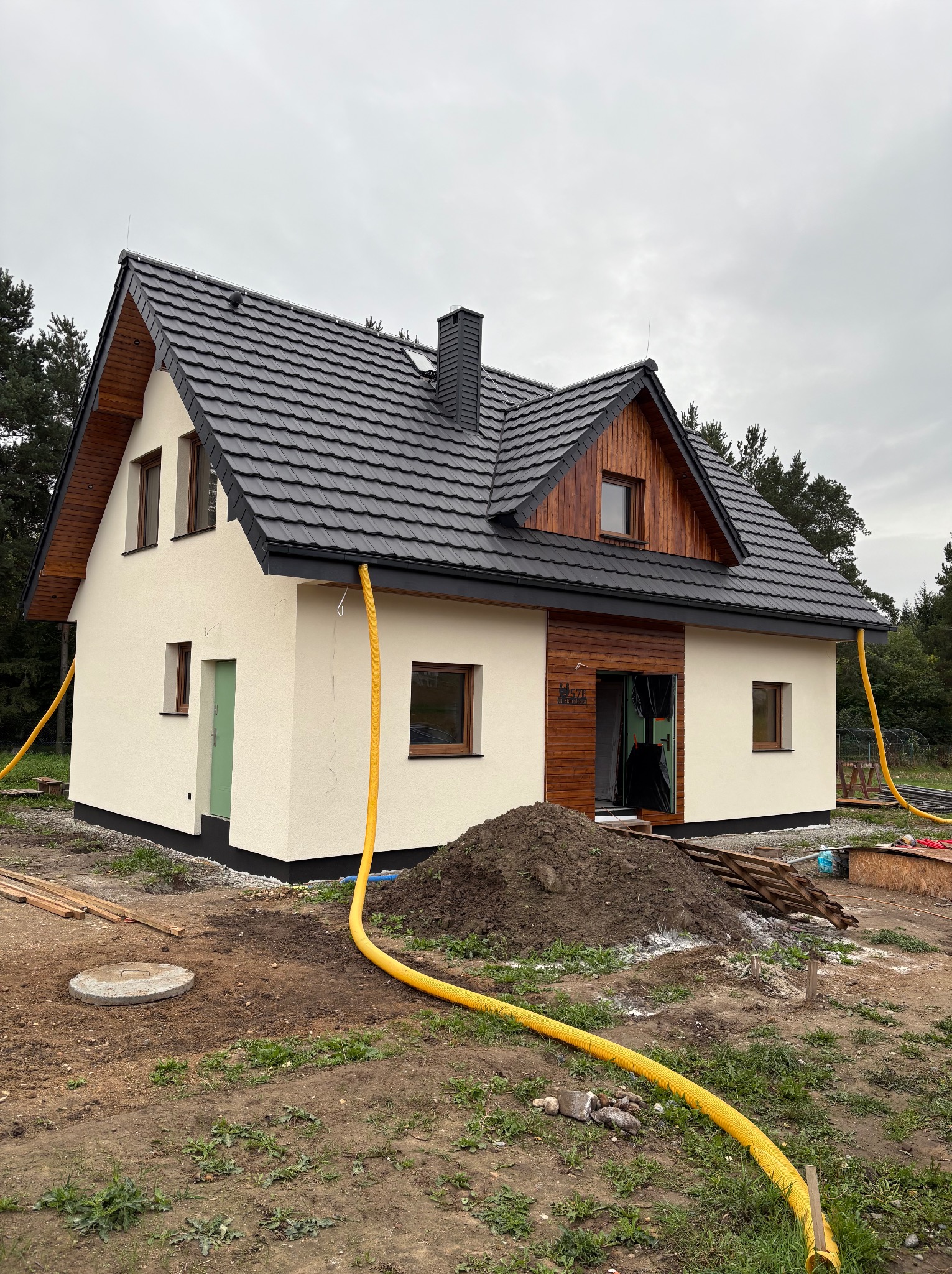 Gotowy dom szkieletowy w Nysie z ciemną dachówką i drewnianymi elementami wykończeniowymi, widoczny z zewnątrz, z żółtymi rurami na trawniku.