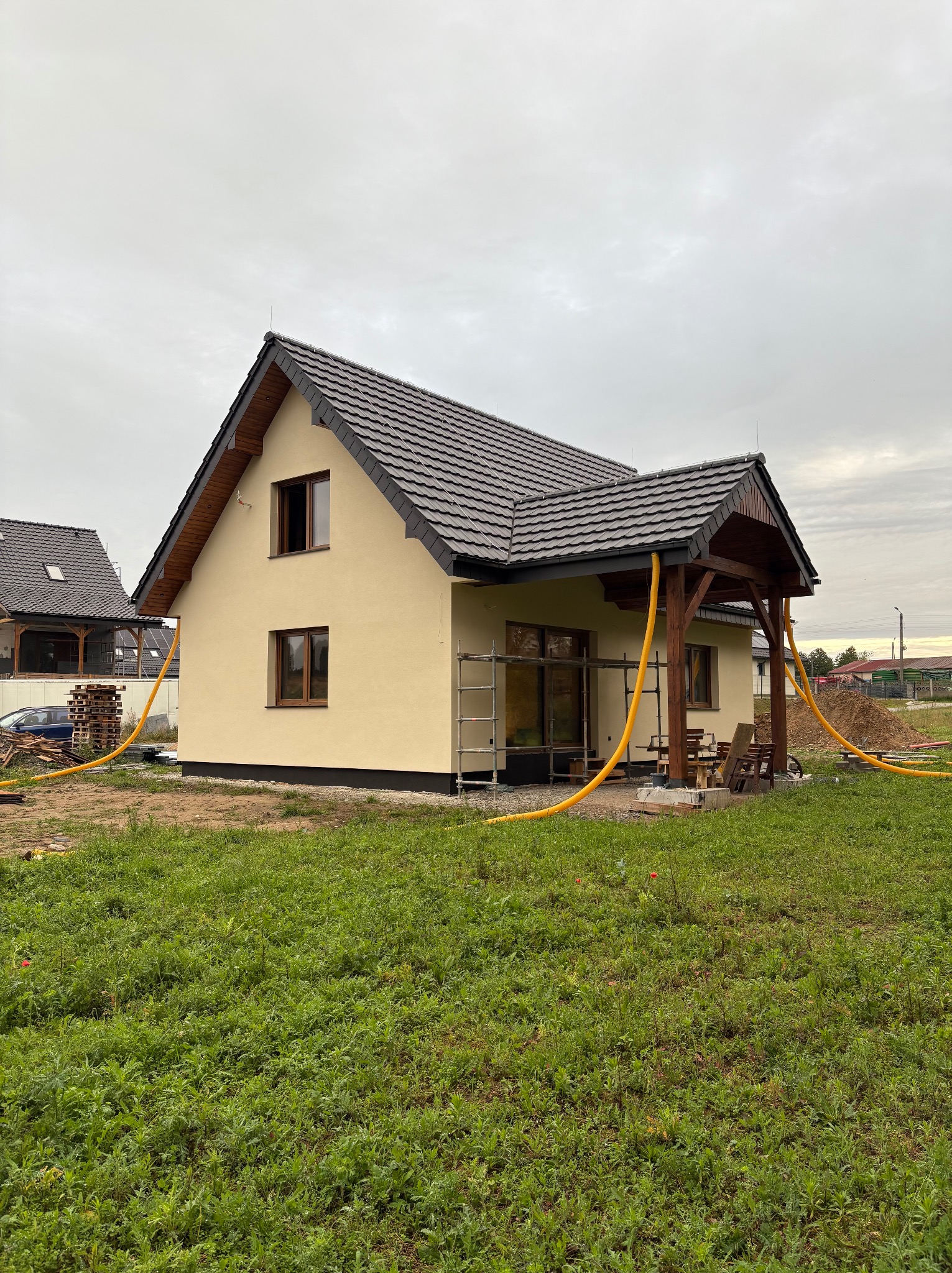 Żółty, otynkowany dom z brązowym dachem, drewnianą werandą i rusztowaniem z boku, otoczony zielonym trawnikiem pod pochmurnym niebem. Widoczne żółte węże.