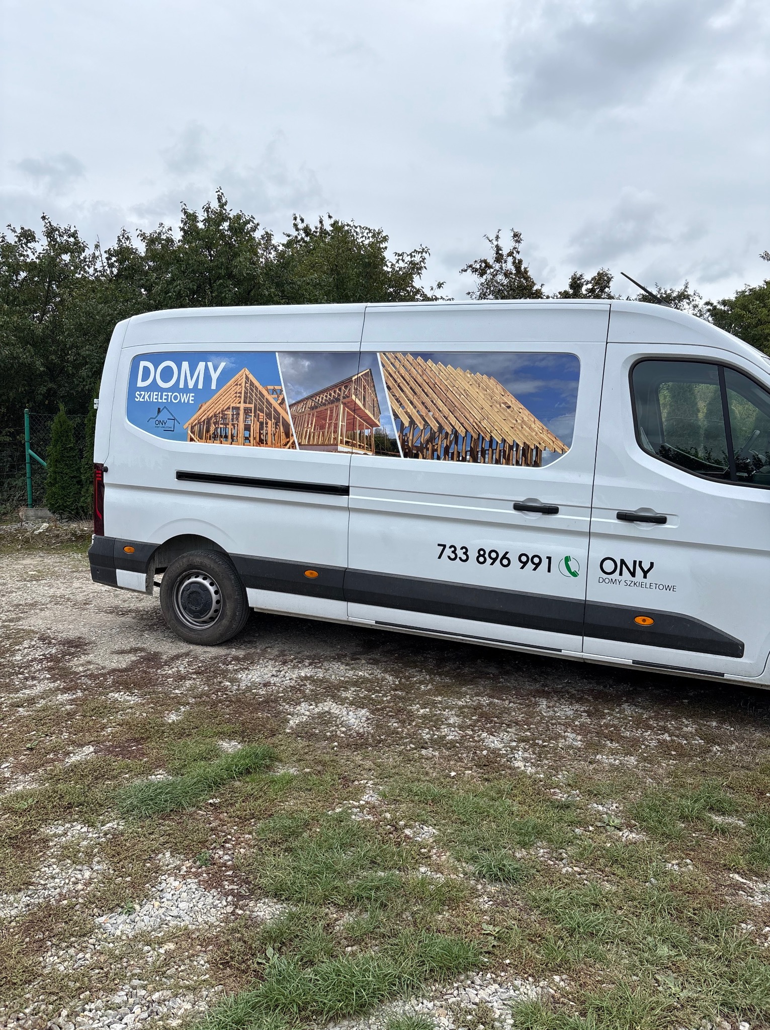 Biały van z reklamą firmy budującej domy szkieletowe, z wizualizacją konstrukcji na boku i numerem telefonu. Tło: drzewa, pochmurne niebo.