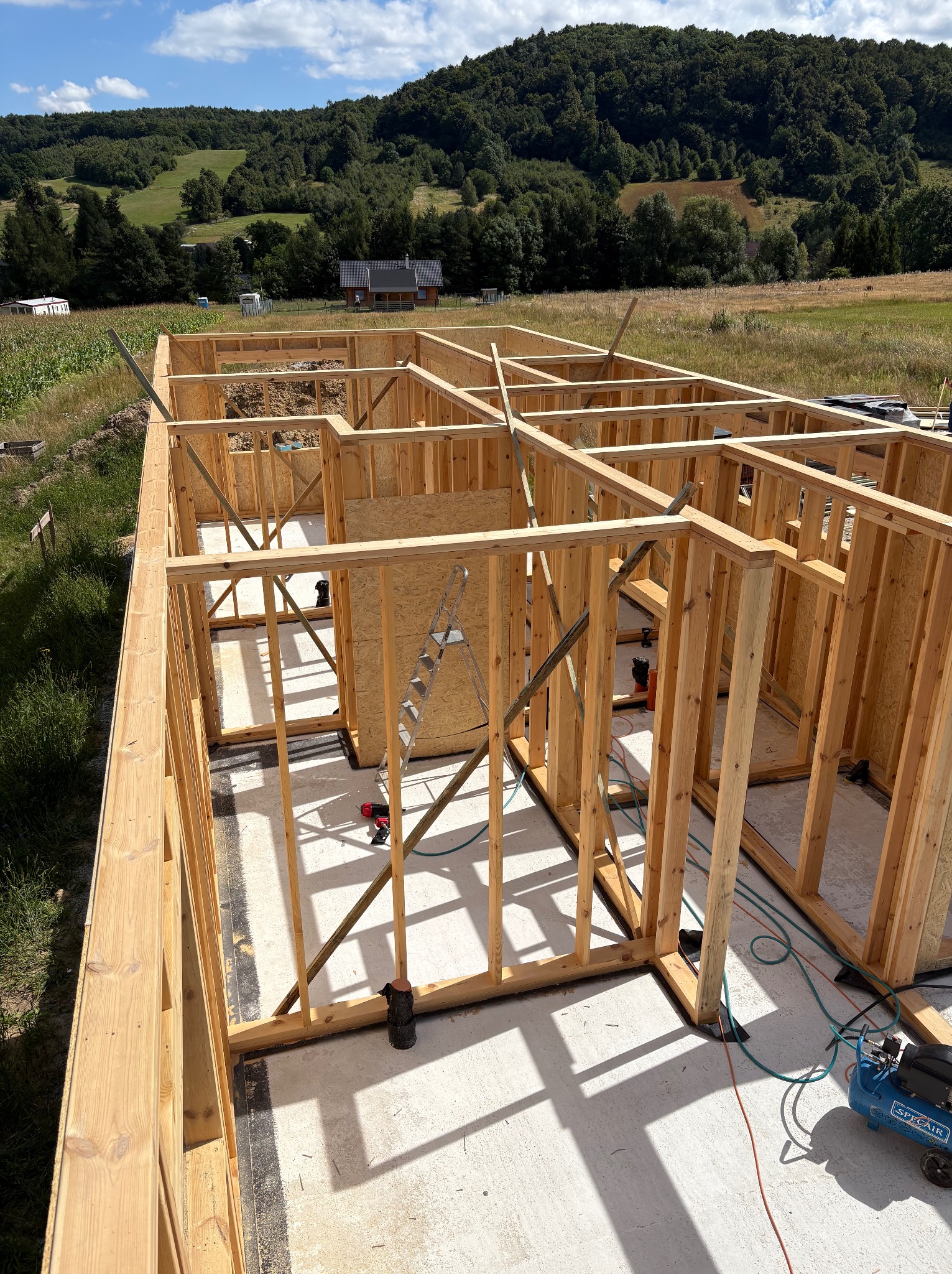 Wnętrze domu szkieletowego w budowie, widok z góry na drewnianą konstrukcję ścian na betonowej płycie fundamentowej, narzędzia pracy, zielone wzgórza w tle.