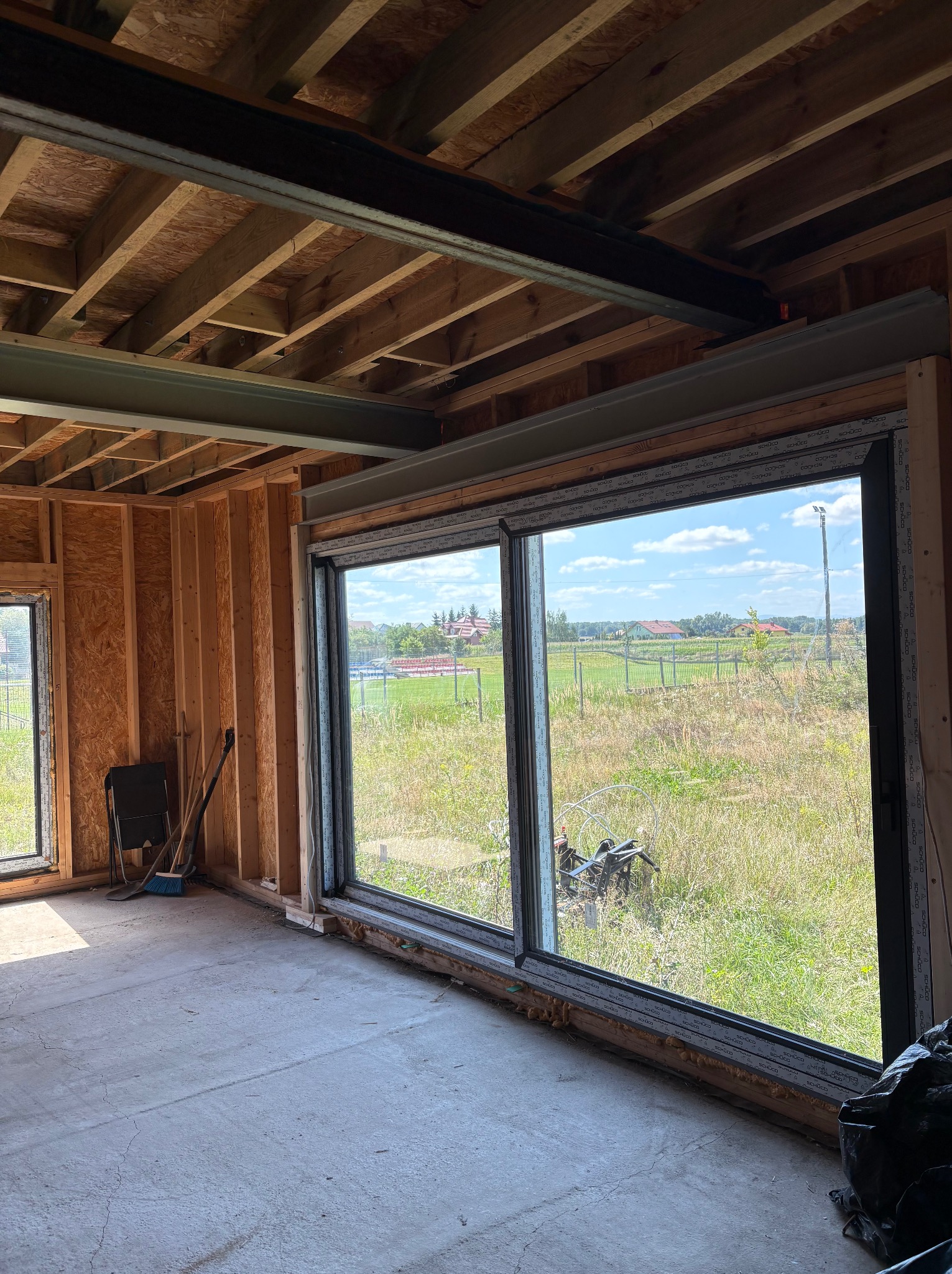 Wnętrze domu w konstrukcji szkieletowej z dużym oknem tarasowym Schüco. Widoczna drewniana konstrukcja ścian, stropu, betonowa posadzka i widok na zewnątrz.