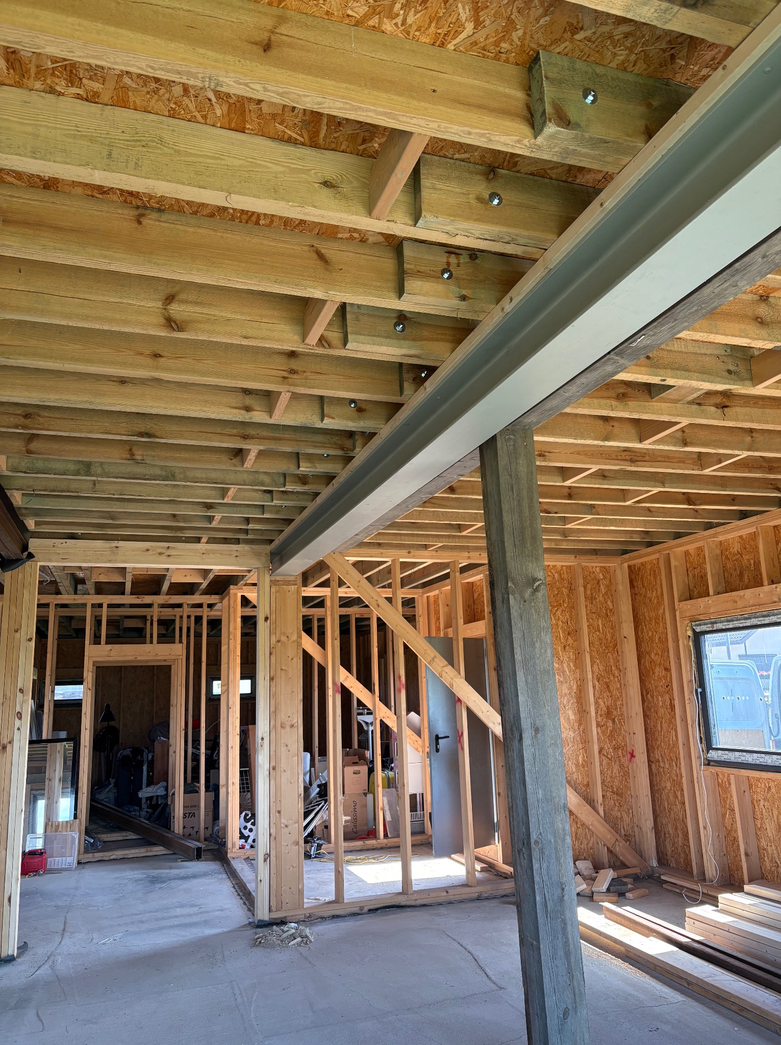 Wnętrze w trakcie budowy domu szkieletowego: drewniana konstrukcja ścian, stropu z widocznymi belkami i metalowym wzmocnieniem. Surowy stan wykończenia.