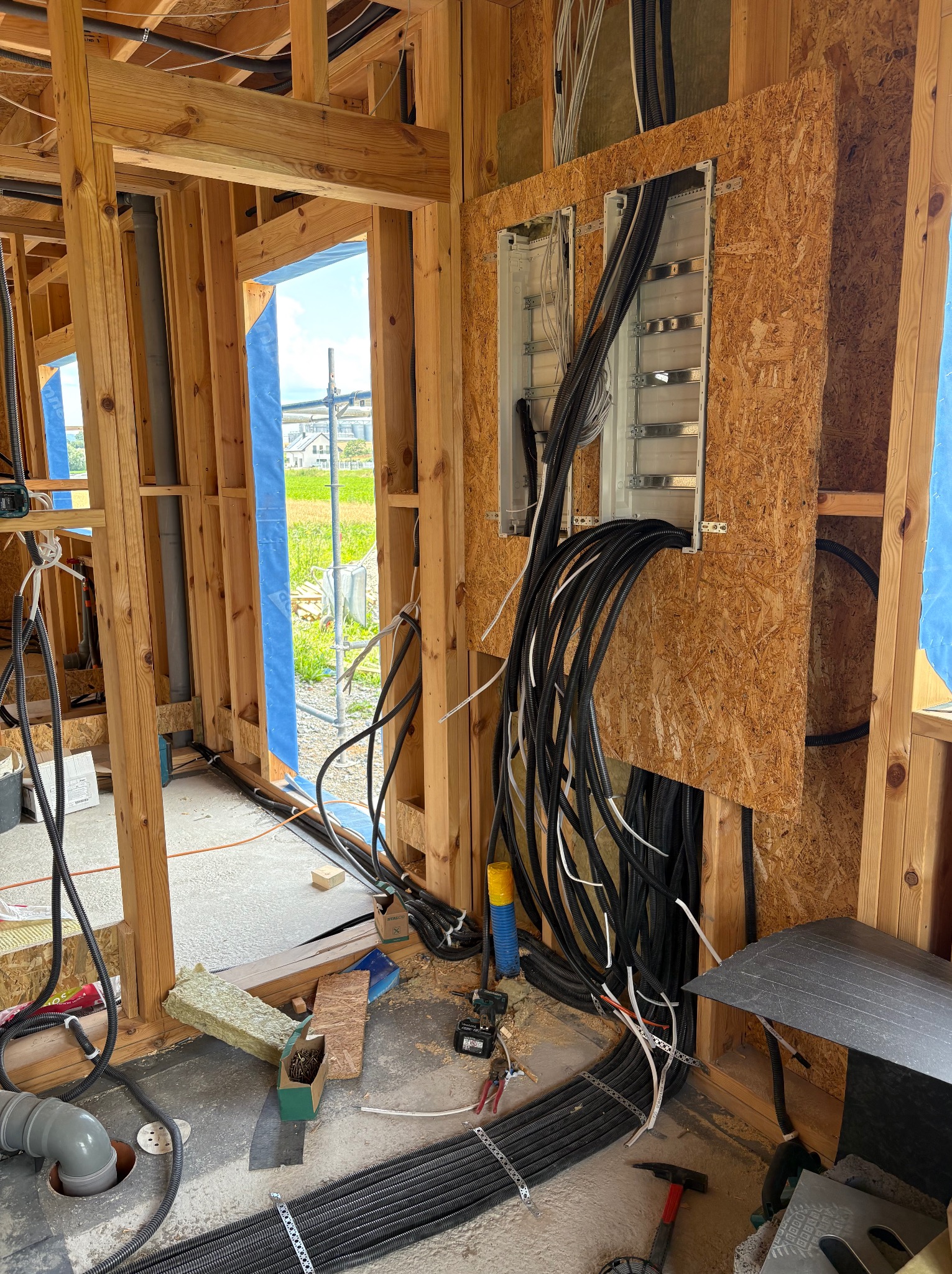 Wnętrze domu w konstrukcji szkieletowej: instalacja elektryczna i rury w ścianach z drewna, widoczne przewody i skrzynki rozdzielcze, fragment widoku na zewnątrz.