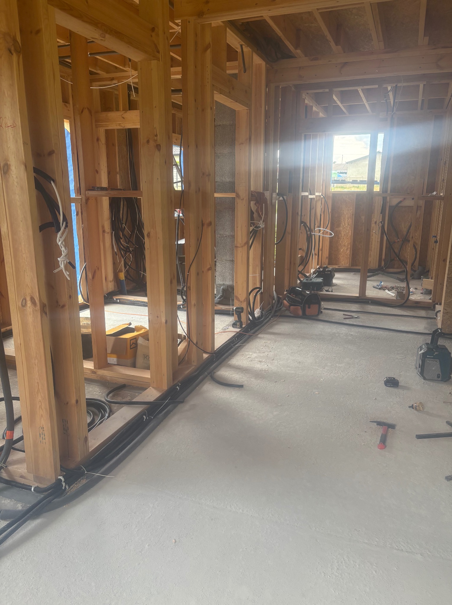 Wnętrze w trakcie budowy: drewniany szkielet domu, instalacja elektryczna prowadzona po podłodze, narzędzia budowlane, widok na okno i dalszą część konstrukcji.