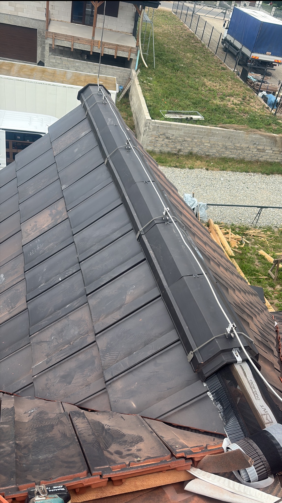 Fragment dachu z ciemnej dachówki ceramicznej, zabezpieczonego linką asekuracyjną, widok z góry na budowę domu w tle.